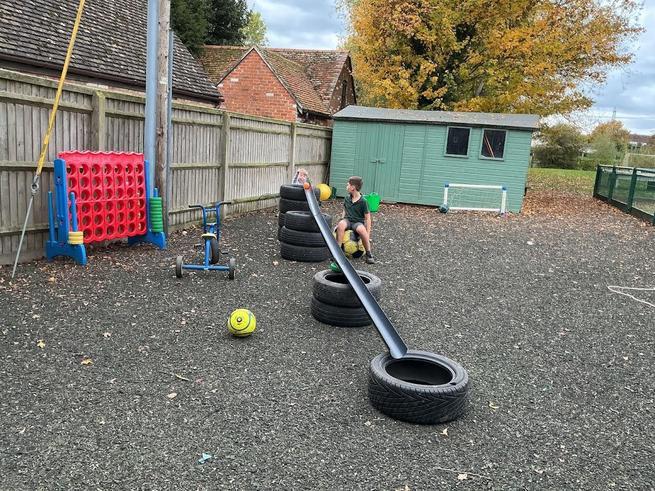 A boy sits on a football in an outdoor playground with Connect Four, a tricycle, a green shed, and autumn trees. Tyres are stacked, and a long ramp extends from them.