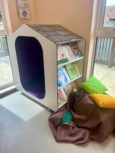 A children's reading nook featuring a white, house-shaped unit with a purple interior, bookshelves filled with books, and colourful cushions on the floor.