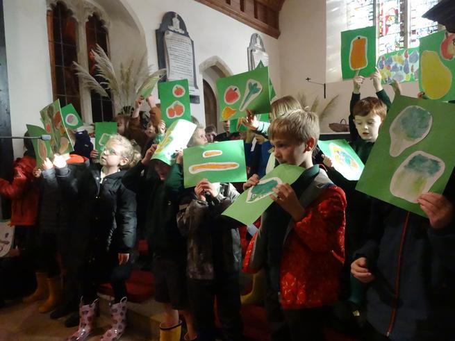 Children in a church, holding green placards with hand-drawn vegetables, likely for a Harvest Festival or play.